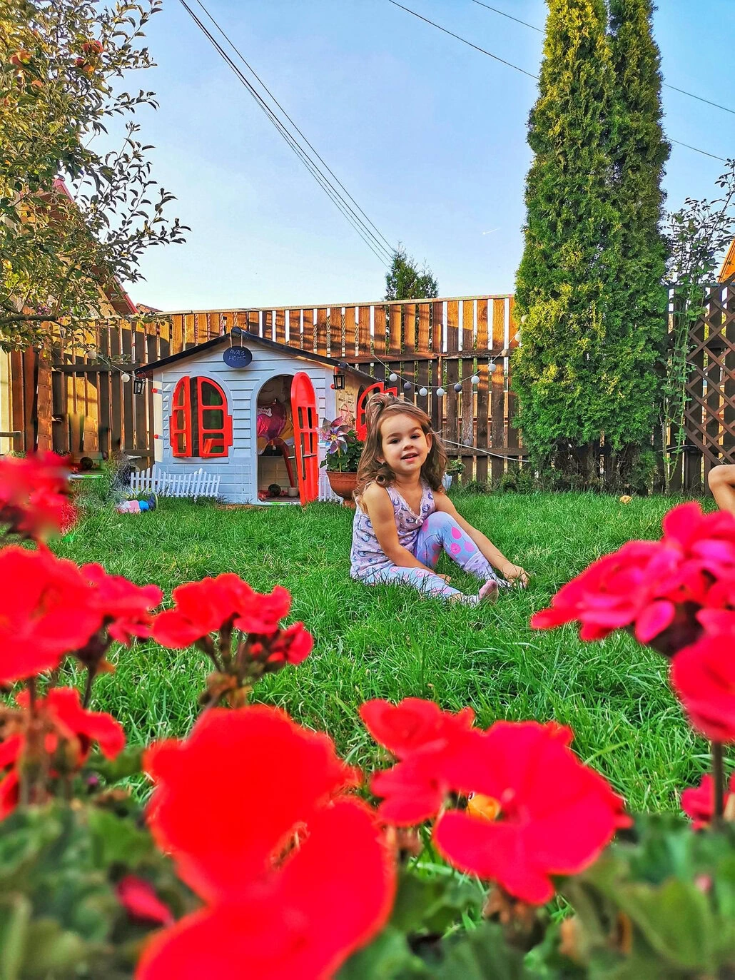 Młoda dziewczyna siedzi na zielonej trawie, uśmiechnięta, na podwórku. Jest blisko kolorowego domku do zabawy z czerwonymi drzwiami i oknami