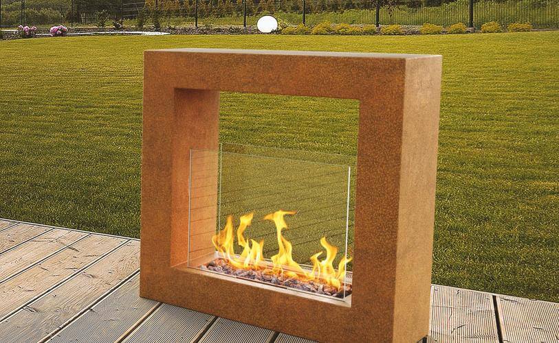 Nowoczesny kominek gazowy zewnętrzny z kwadratową ramą i widocznymi płomieniami stoi na drewnianym tarasie. Za tarasem znajduje się dobrze utrzymany trawnik i ogród, otoczony czarnym metalowym płotem. W tle żywopłot i zieleń. 