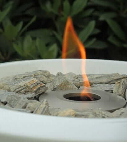 Małe palenisko na świeżym powietrzu z pojedynczym płomieniem wydobywającym się z centralnego okrągłego otworu otoczonego szarymi kamieniami. W tle znajdują się zielone rośliny liściaste. 