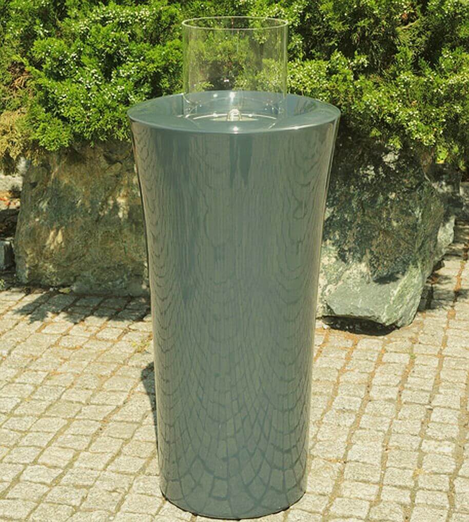 Nowoczesny, cylindryczny biokominek ogrodowy z eleganckim, metalicznym wykończeniem stoi na kamiennej powierzchni. Na górze zamontowany jest przezroczysty pojemnik na popiół. Tło składa się z zieleni i dużych skał. 