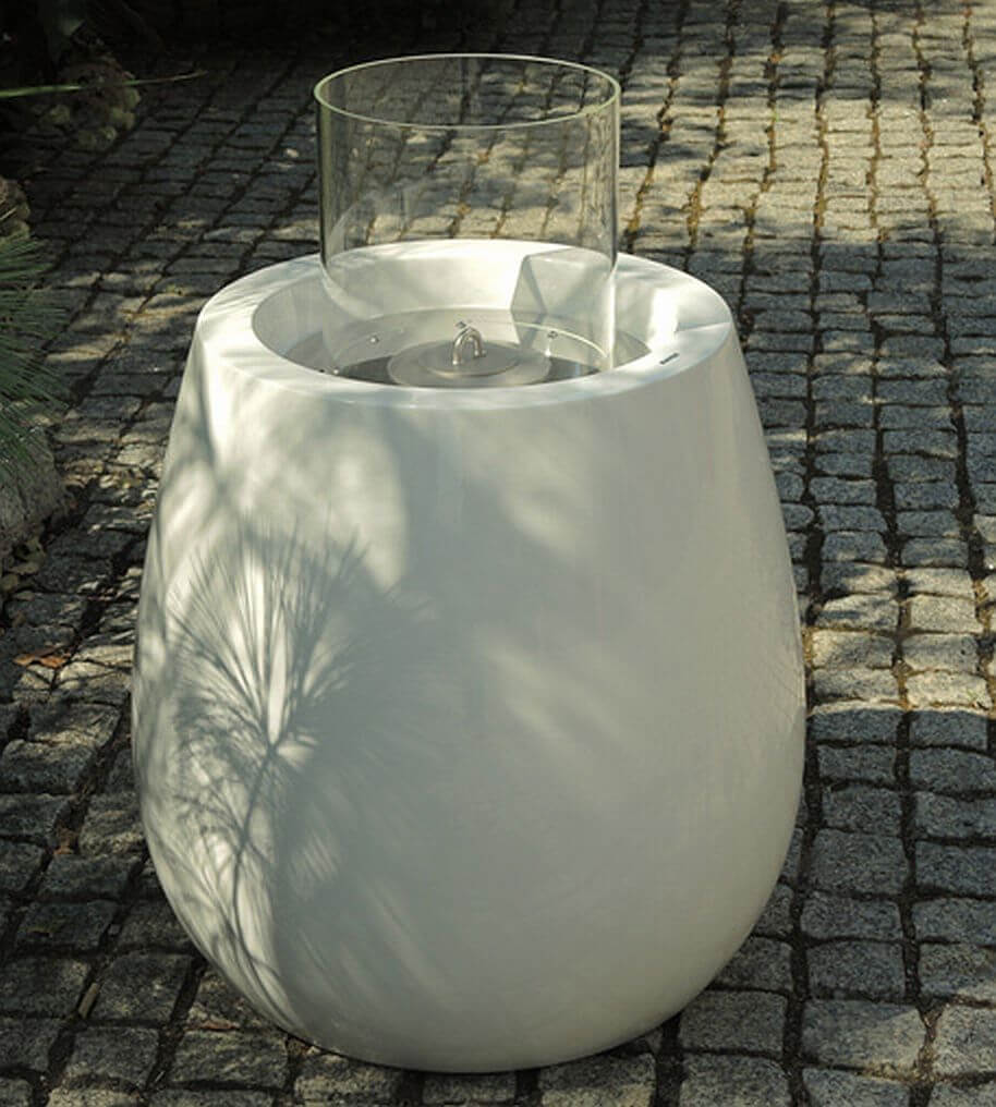 Nowoczesny biały kominek zewnętrzny ze szklaną obudową na brukowanej powierzchni, rzucający miękkie cienie z pobliskich drzew. Elegancka konstrukcja ma zaokrągloną podstawę i cylindryczny blat, na którym widoczny jest mały płomień. 