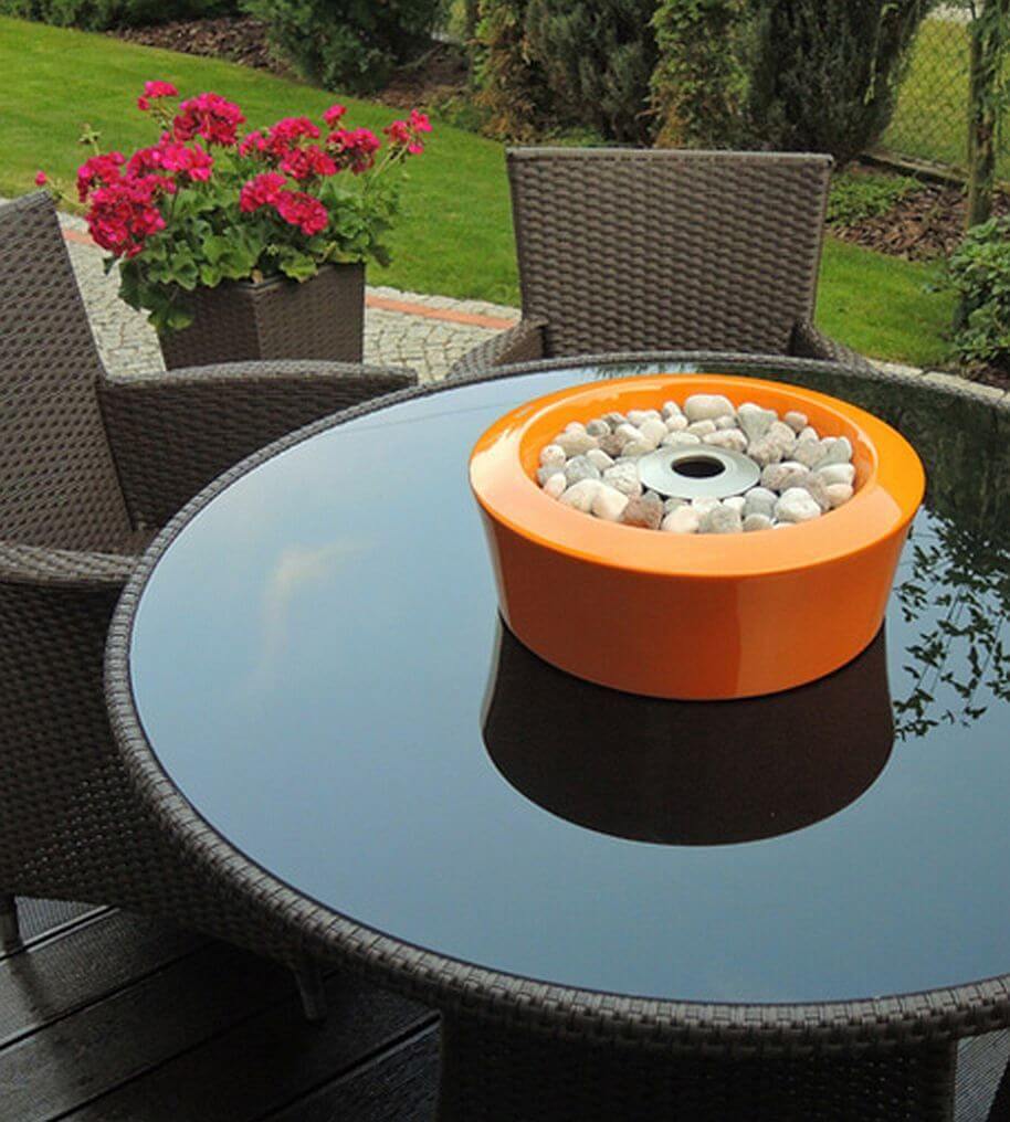 Nowoczesne patio na świeżym powietrzu z okrągłym stołem ze szklanym blatem otoczonym wiklinowymi krzesłami. Na stole stoi pomarańczowa okrągła donica wypełniona kamieniami. W tle doniczka z żywymi czerwonymi kwiatami stoi na drewnianym tarasie obok zielonego trawnika. 