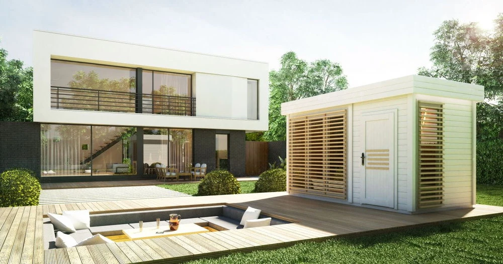Nowoczesna, biała sauna AZALIA z dużymi oknami, drewnianym tarasem, zagłębioną częścią wypoczynkową na świeżym powietrzu i małym białym budynkiem ogrodowym; bujna zielona trawa i drzewa otaczają nieruchomość.