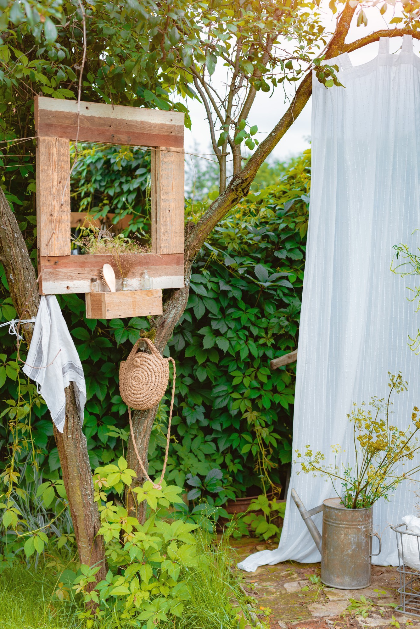 Scena z prysznicem na świeżym powietrzu przedstawia drzewo o rustykalnej drewnianej konstrukcji, w którym znajdują się niezbędne akcesoria prysznicowe. Obok wisi biała zasłona zapewniająca prywatność. W pobliżu wisi słomkowy kapelusz i ręcznik, a na nim rośliny, konewka i stylowe drewniane lustro