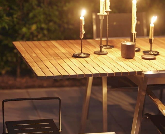 Drewniany stół do jadalni na świeżym powietrzu zastawiony jest zapalonymi świecami w metalowych świecznikach, tworząc ciepłą i przytulną atmosferę. Stół otoczony jest krzesłami, a na rozmytym tle widać zieleń, wskazującą na wieczorną oprawę.