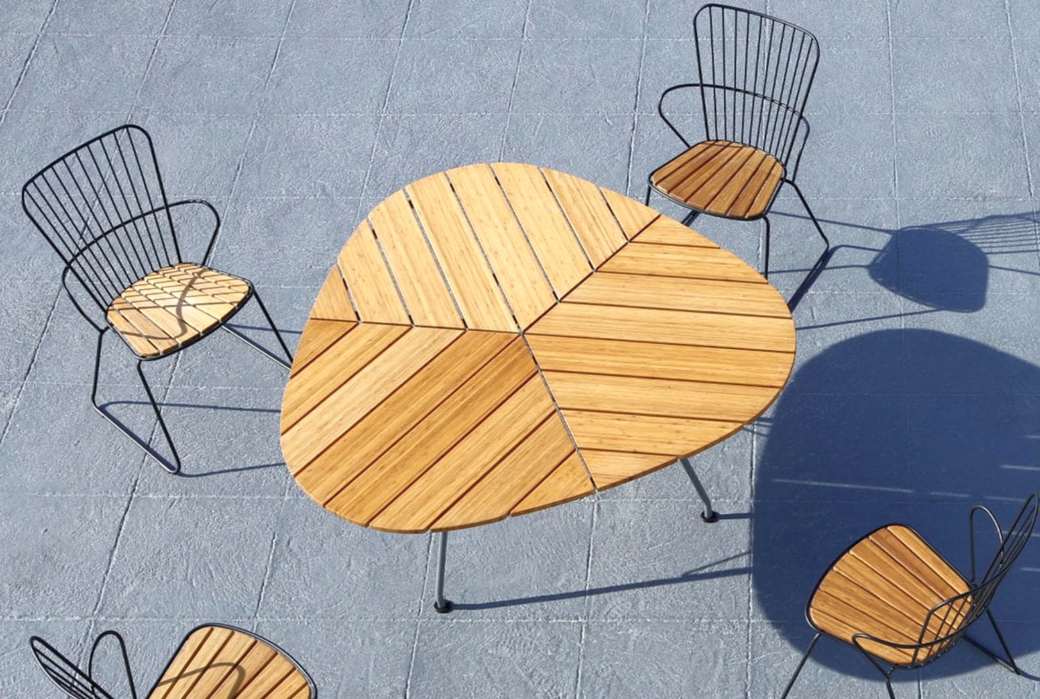 Zdjęcie z góry przedstawiające unikalny trójkątny drewniany stół z trzema segmentami i czterema pasującymi krzesłami. Stół i krzesła mają metalowe ramy, a siedziska i blat wykonane są z drewna. Są one umieszczone na szarej, teksturowanej powierzchni zewnętrznej.