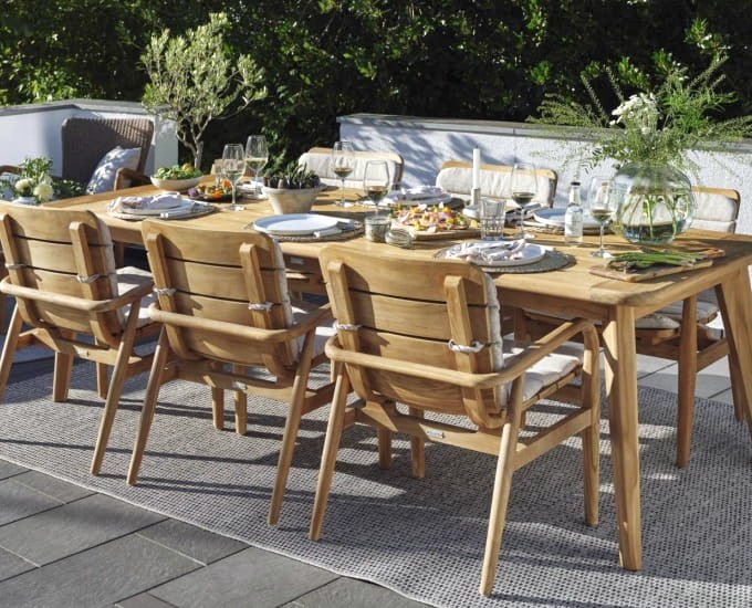 Do posiłku nakryty jest duży drewniany stół jadalny, a wokół niego starannie ustawiono osiem krzeseł. Talerze, szklanki, sztućce i różne naczynia ustawiane są na stole, na którym znajdują się również kompozycje kwiatowe w wazonach. Akcja rozgrywa się na patio z zielenią w tle.