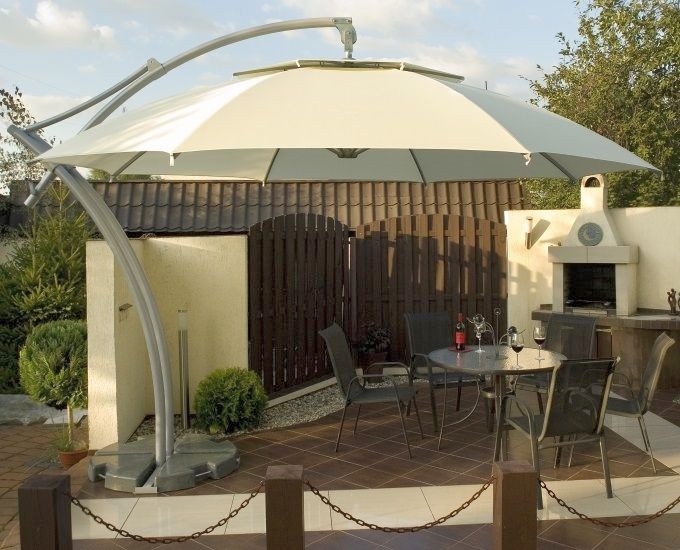 Scena na patio z okrągłym szklanym stołem otoczonym czterema metalowymi krzesłami. Na stole stoi otwarta butelka wina, dwa kieliszki do wina i czerwona świeca. Duży, biały parasol wspornikowy ocienia okolicę. Za domem drewniany płot i kuchnia na świeżym powietrzu z grillem.