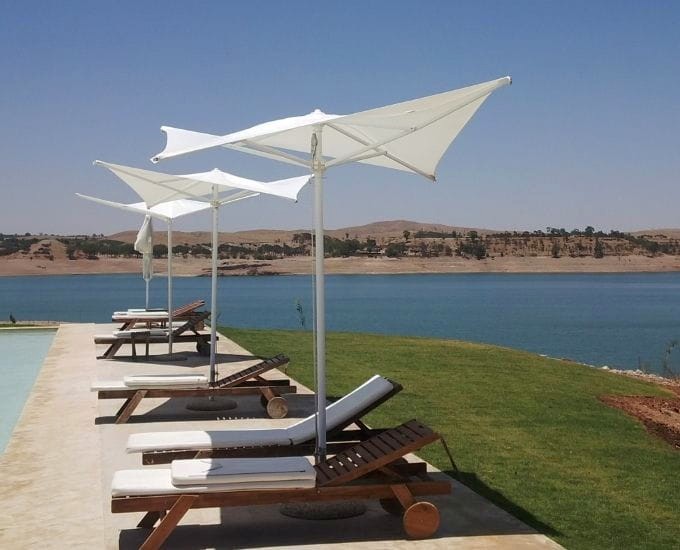 Spokojny teren przy basenie z kilkoma drewnianymi leżakami, każdy w połączeniu z białym parasolem słonecznym. Z basenu i leżaków roztacza się widok na duży zbiornik wodny z pagórkowatym krajobrazem w tle pod czystym, błękitnym niebem.