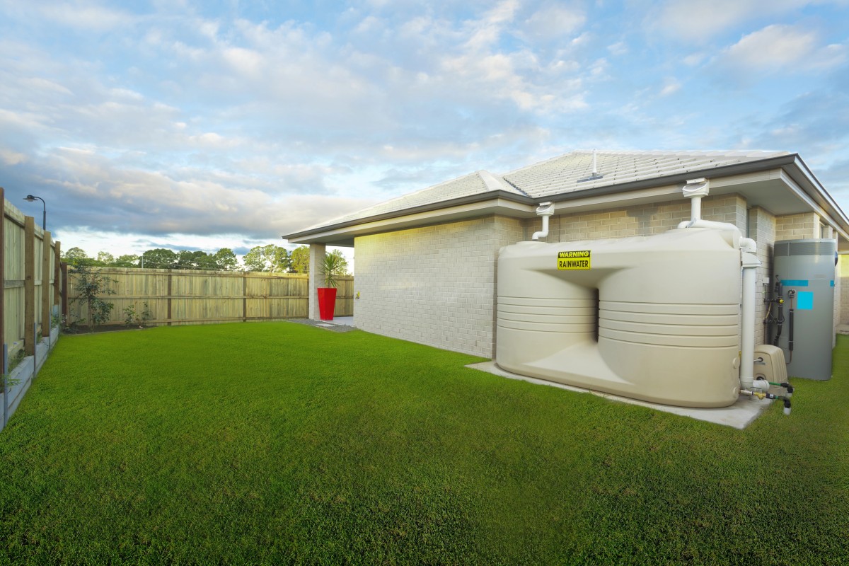 Podwórko z zielonym trawnikiem i białym domem po prawej stronie. Do domu dołączony jest duży, beżowy zbiornik na wodę z napisem „Zbiornik na deszczówkę”. Obok zbiornika znajduje się niebieski system filtrów. W tle obok drewnianego płotu stoi wysoka, czerwona donica. 