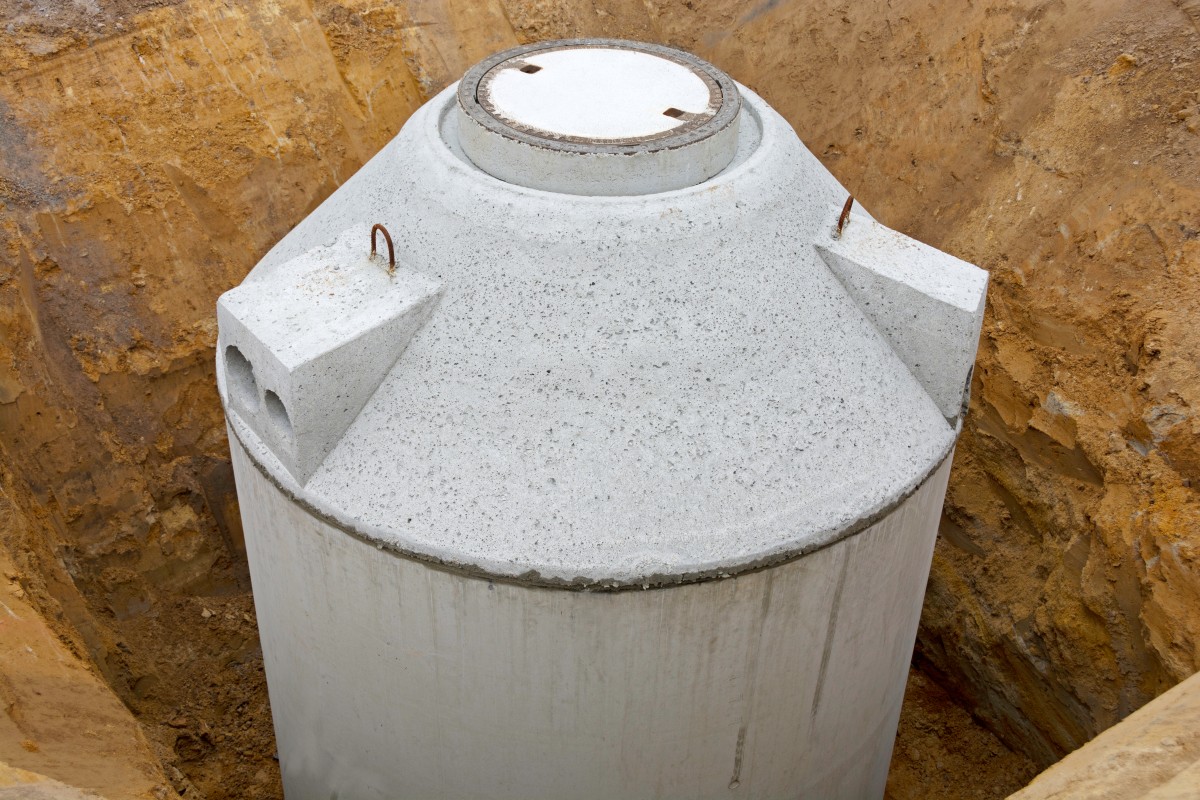 Duże betonowe szambo częściowo zakopane w ziemi, z okrągłą pokrywą dostępową na górze. W okolicy widoczna jest odsłonięta gleba, co wskazuje na trwające prace budowlane lub instalacyjne. 