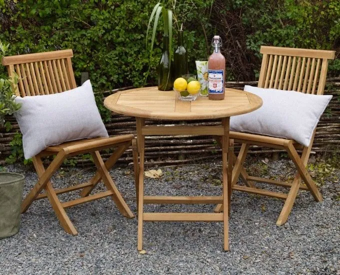 Mały, okrągły drewniany stolik z dwoma drewnianymi krzesłami ustawiony na zewnątrz na żwirowej powierzchni. Na stoliku stoi butelka, dwie cytryny i wazon z zielenią. Oba krzesła mają białe poduszki. W tle widać bujną zieloną roślinność i pleciony drewniany płot.