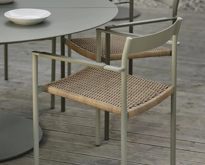 Minimalistyczny zestaw ogrodowy na patio z okrągłym metalowym stołem i pasującymi krzesłami. Krzesła mają plecione siedziska i eleganckie podłokietniki. Stół ma płytką miskę, a zestaw znajduje się na drewnianym tarasie. Paleta kolorów jest neutralna i spójna.