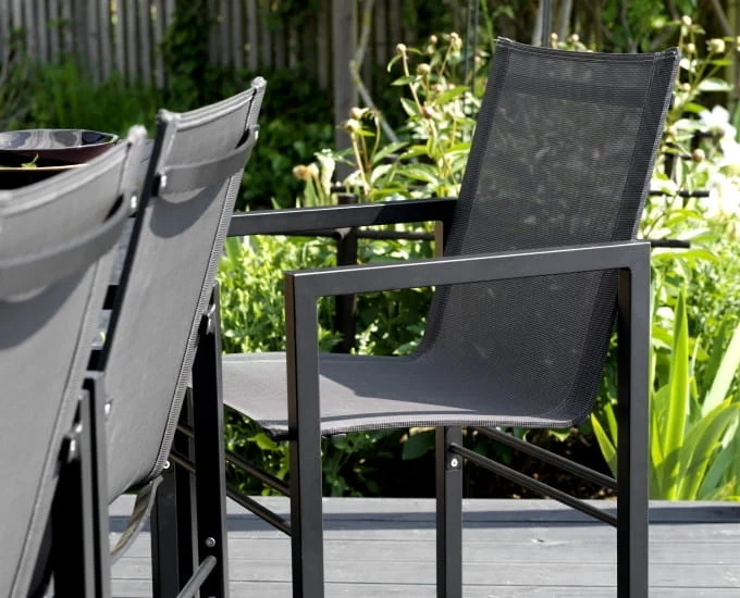 Zestaw nowoczesnych czarnych krzeseł ogrodowych otaczających stół na patio. Krzesła mają oparcia z siatki i siedziska z metalowymi ramami. Tło zdobią bujne zielone rośliny i drewniany płot.