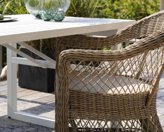 Krzesło wiklinowe z beżową poduszką ustawione obok białego stolika ogrodowego. Na stoliku znajdują się szklane elementy dekoracyjne i przezroczysty wazon. Tło przedstawia zielone rośliny, sugerujące ustawienie w ogrodzie lub na patio.