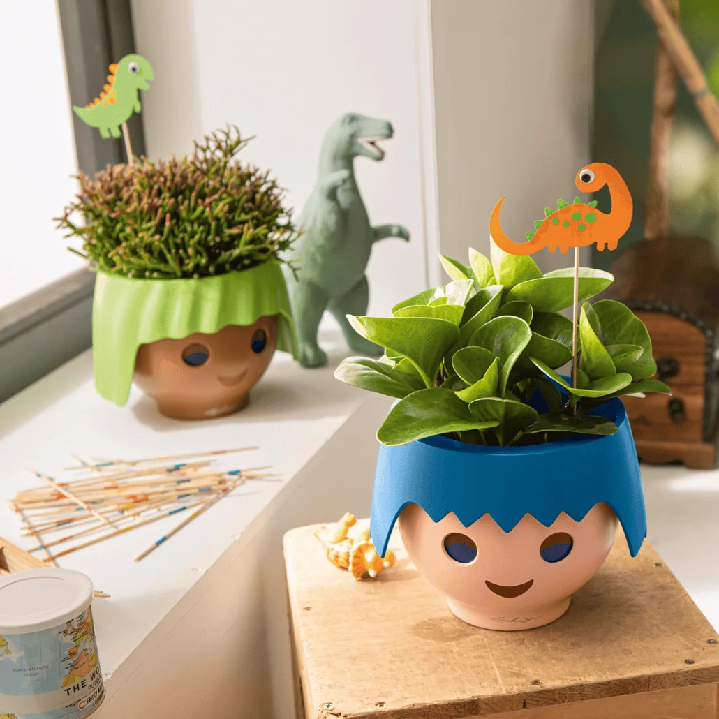 Dwie zabawne doniczki w kształcie uśmiechniętych twarzy stoją na parapecie. Jedna z nich ma zielone "włosy" z trawą, a druga niebieskie "włosy" z roślinami liściastymi. Obie mają dekoracje z dinozaurami z kreskówek wśród roślin.