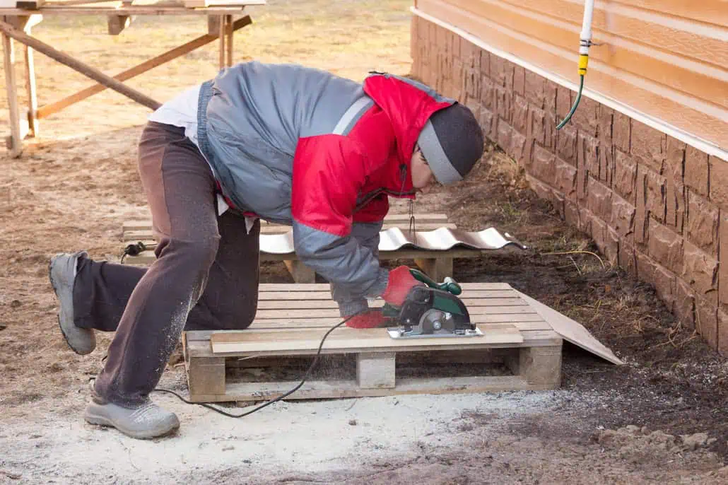 Osoba w czerwonej kurtce i kapeluszu klęczy na drewnianej palecie, fachowo używając piły tarczowej do jej cięcia. Pracują na zewnątrz w pobliżu budynku z sidingiem i kamiennym fundamentem.