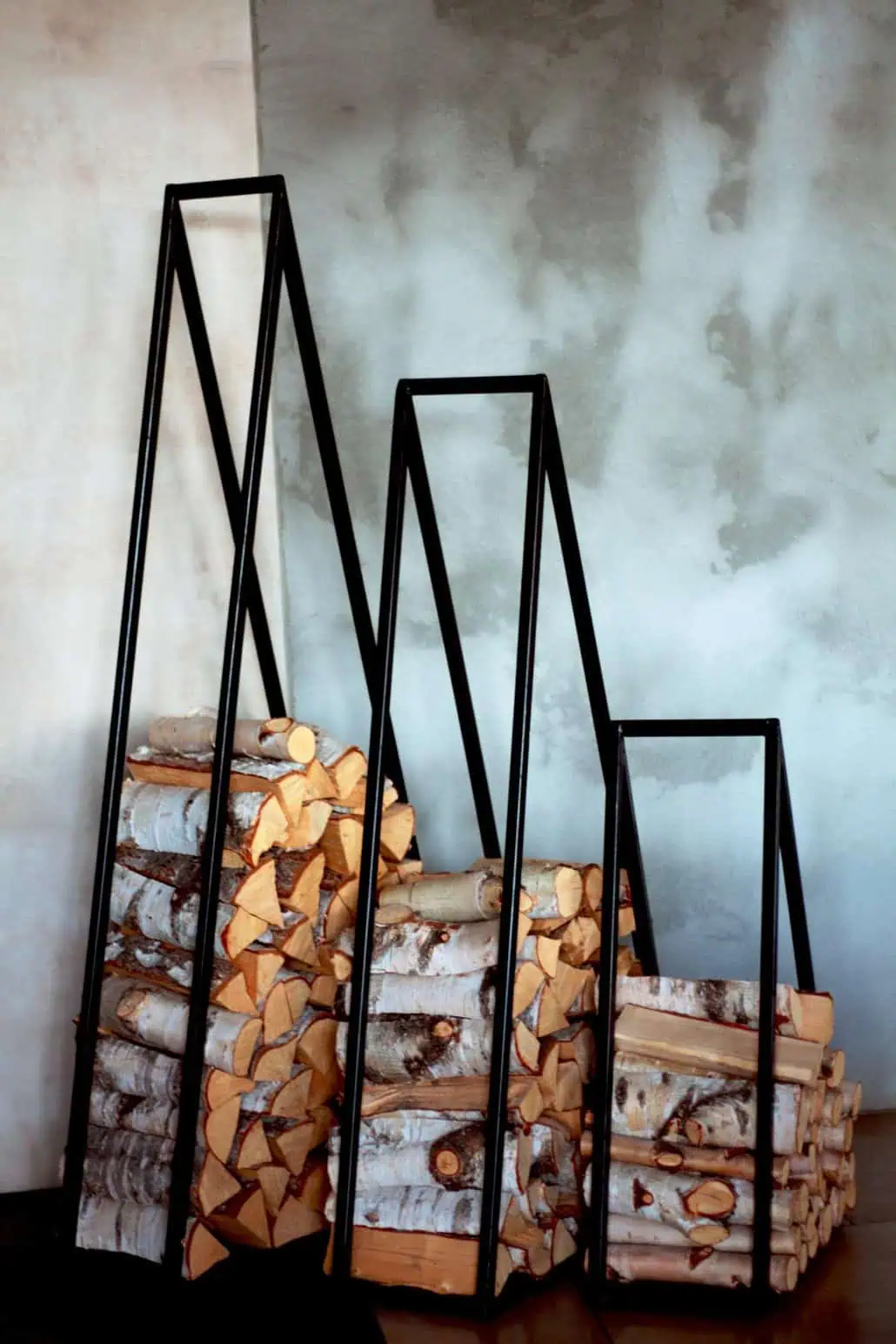 Nowoczesne metalowe uchwyty na drewno opałowe ze stosami drewna opałowego na tle szarej ściany tworzą uderzającą geometryczną ekspozycję. Trójkątne uchwyty różnią się wysokością, a starannie ułożone drewno do ognia eksponuje swoją naturalną korę i cięte powierzchnie.