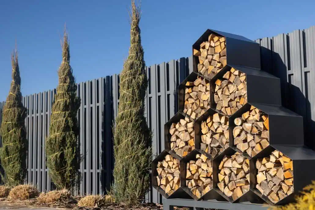 Starannie ułożony stos drewna do palenia jest ułożony w sześciokątnych przegrodach przy wysokim, ciemnoszarym płocie. Wysokie, przycięte drzewa iglaste, w tym tuja owinięta, rosną wzdłuż płotu pod czystym, błękitnym niebem.