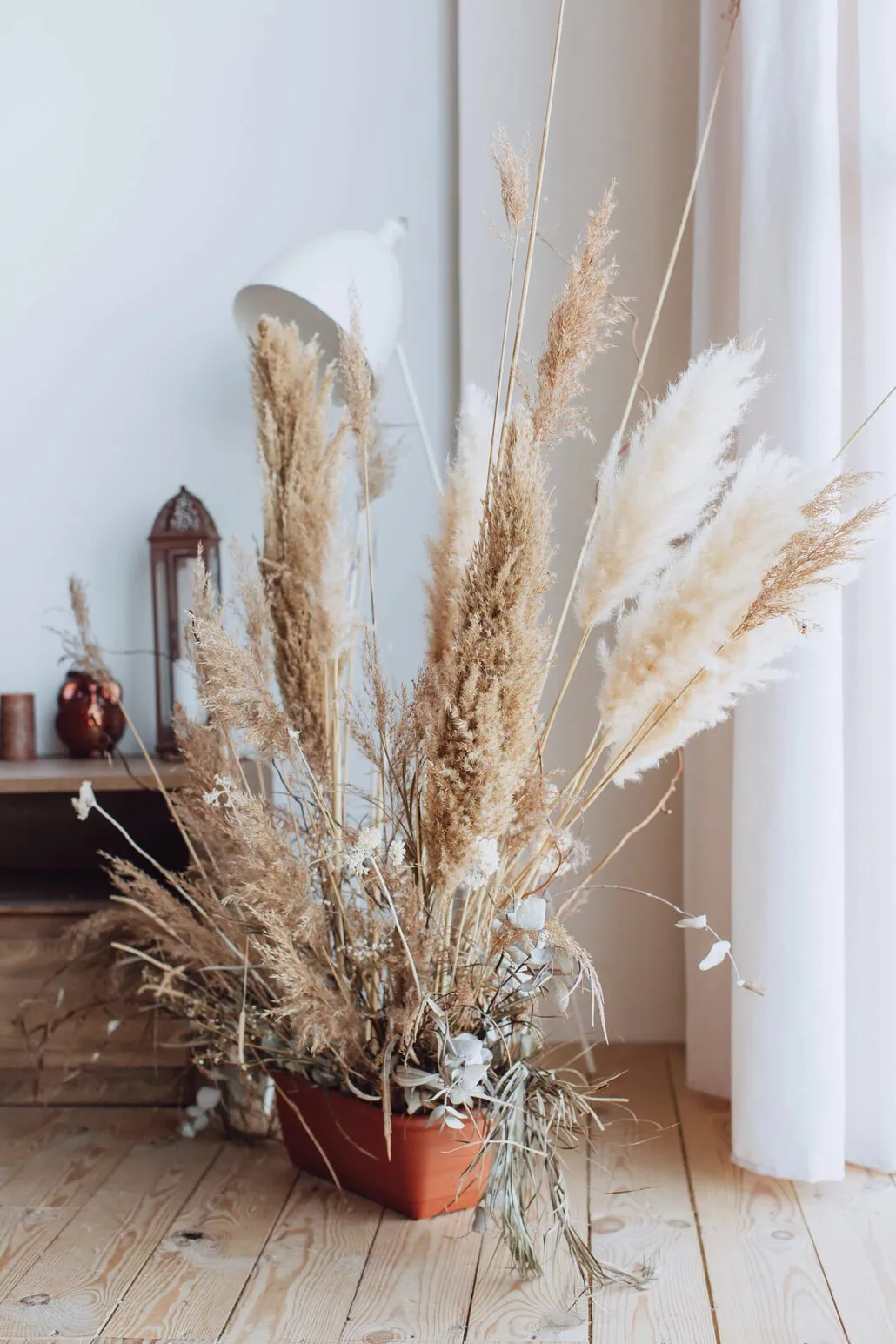 Terakotowa donica wypełniona wysokimi trawy Suszone i różnymi suszonymi liśćmi stoi na drewnianej podłodze w pobliżu okna z białymi zasłonami, w delikatnie oświetlonym, minimalistycznym pokoju.