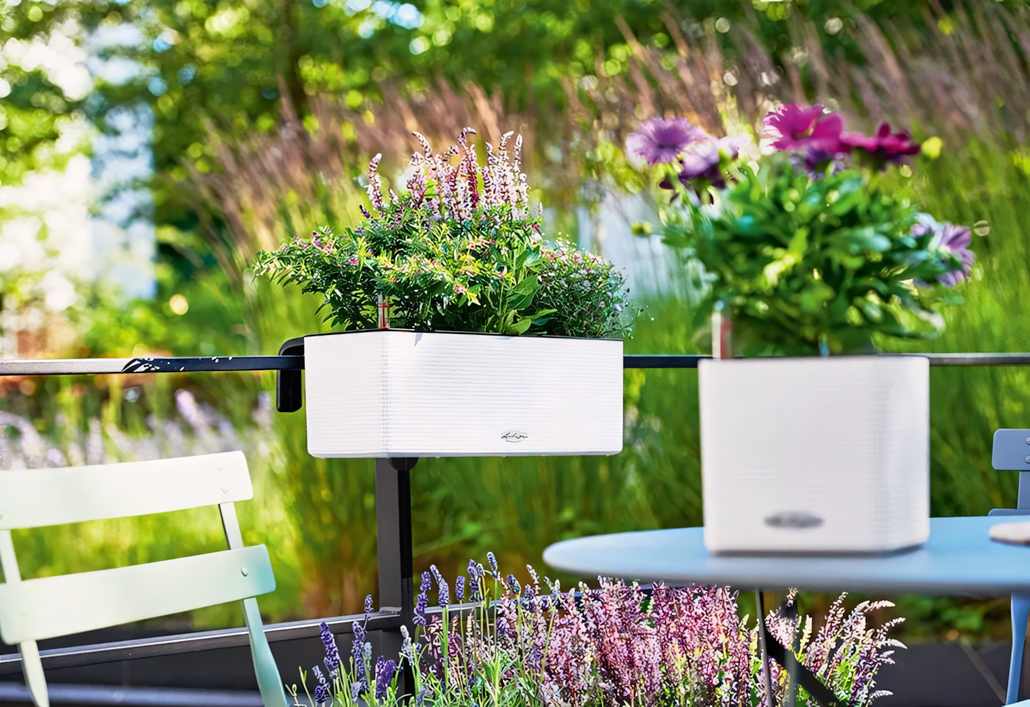 Białe donice z kwitnącymi fioletowymi i zielonymi kwiatami są przymocowane do czarnej poręczy na patio na zewnątrz. W tle widać jasnozielone krzesło i bujną zieleń.