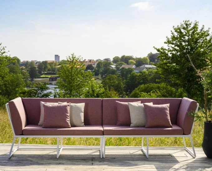Ażurowa 4-osobowa sofa „Vence” z różowymi poduszkami i białą ramą na drewnianym tarasie z widokiem na park.