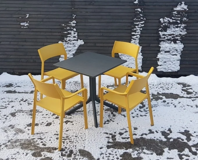 Kwadratowy stolik „Clip Durel” o wymiarach 70 x 70 z czterema żółtymi krzesłami na zaśnieżonym patio.