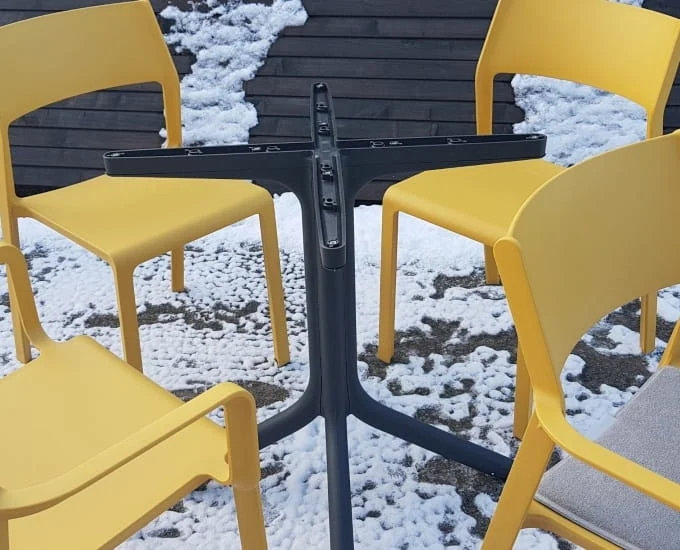 Ciemnoszara podstawa stolika „Clip” z krzyżową podstawą w otoczeniu czterech żółtych krzeseł na ośnieżonym podłożu.