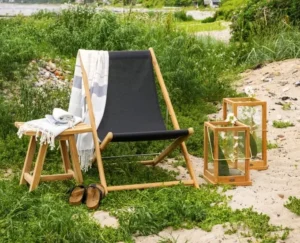 Składany drewniany leżak Sindos z czarnym siedziskiem, stolikiem i dwoma lampionami na piaszczystej plaży.