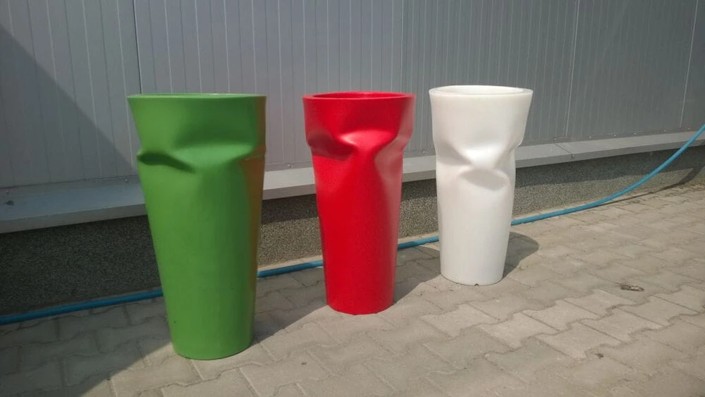 Trzy wysokie, cylindryczne donice „Pogięta Bent” z wgłębieniami w kolorach zielonym, czerwonym i białym, ustawione w rzędzie na bruku.