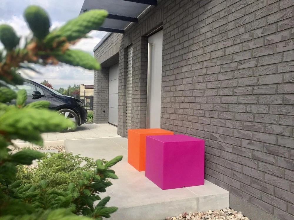 Pomarańczowe i magenta donice „Kostka Kubik SQUARE” na betonowym patio przy szarej ceglanej ścianie.