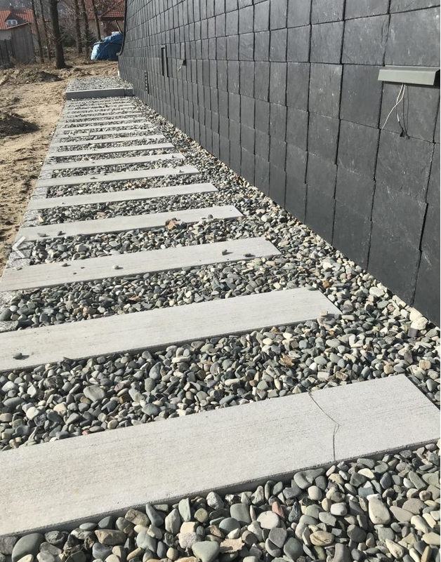 Płyta chodnikowa o wymiarach 25 x 120 z betonu architektonicznego, ułożona w ścieżkę na żwirze przy płytkach ściennych.