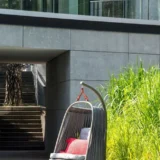 Szary fotel wiszący „Huśtawka Cocoon Art Deco” z czerwoną poduszką, umieszczony na stelażu na drewnianym tarasie przy wysokiej trawie.