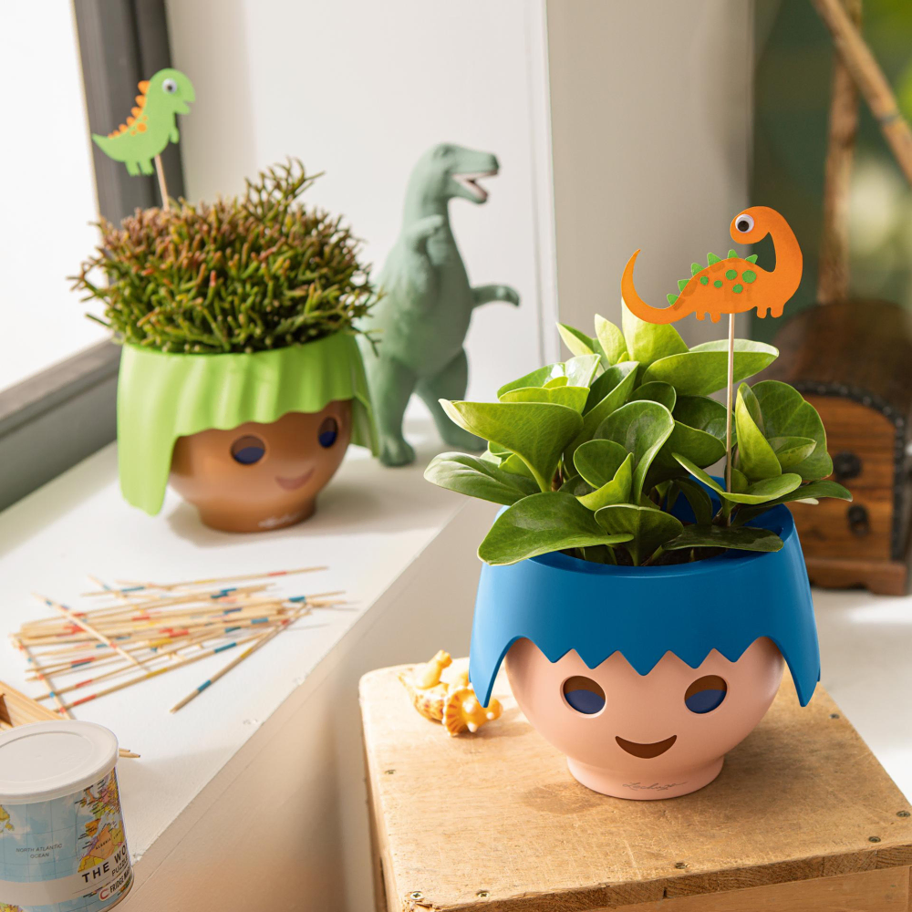 Zielona i niebieska doniczka OJO do pokoju dziecięcego Lechuza&Playmobil w kształcie głowy z roślinami przypominającymi dinozaury.