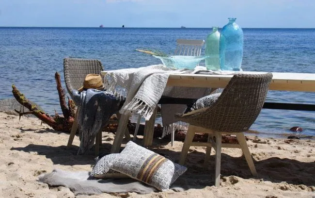 Dwa krzesła Clifton New na drewnianych nogach przy plażowym stole, wazony, koc i poduszki.