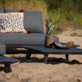 Ciemnoszara, niska sofa modułowa Bart dla dwóch osób, wzorzysta poduszka, niski stolik, ananas i miska na piaszczystym podłożu.