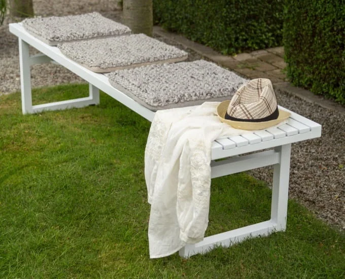Aluminiowa ławka „Kordoba” z trzema szarymi poduszkami, białą tkaniną i kapeluszem typu fedora na trawniku.