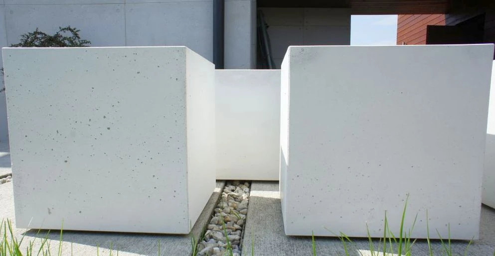 Dwie kwadratowe, białe donice z betonu kostkowego o wymiarach 80 x 80 x 80 cm na wybrukowanym tarasie, z przerwą wypełnioną żwirem.