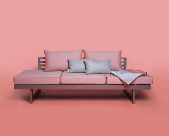 Różowa sofa Stern New Holly z oparciem z listewek, dwiema poduszkami, dwiema szarymi poduszkami i szarym kocem.