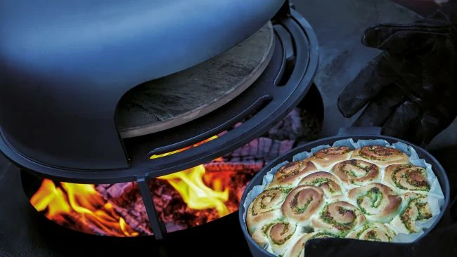 Ręce w rękawiczkach trzymają blachę z bułeczkami ziołowymi obok pieca. Ogródowy piec do pizzy z kamiennym dnem i ogniem.