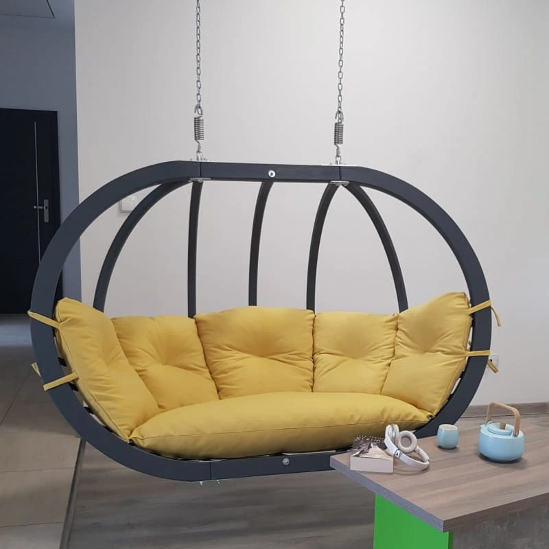 Ciemnoszary dwuosobowy kosz wiszący O-Zone Premier Swing Pod z żółtymi poduszkami, obok stolika z czajnikiem i słuchawkami.