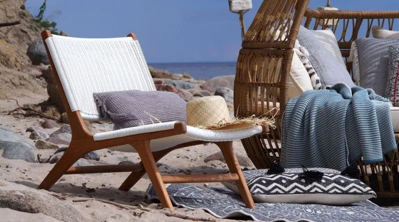 Biały fotel ogrodowy Tobago z drewna tekowego z plecionym siedziskiem, poduszkami i słomkowym kapeluszem na plaży.