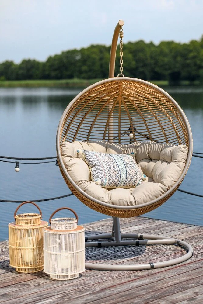 Huśtawka koszowa Cocoon De Luxe, wiszące krzesło typu kokon z beżowymi poduszkami i lampionami na drewnianym tarasie nad jeziorem.