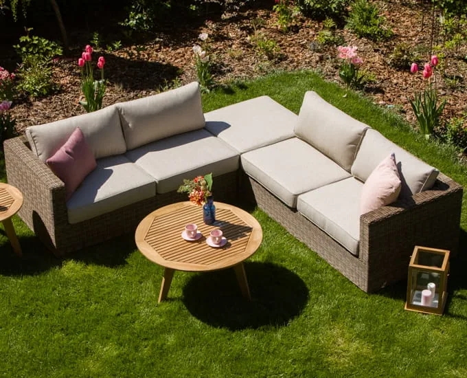 Modułowa sofa narożna Le Mans w kształcie litery L z jasnymi poduszkami i okrągłym drewnianym stolikiem kawowym na trawniku.