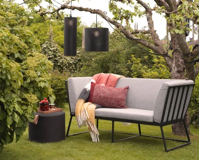 Ażurowa dwuosobowa sofa „Vence”, szare poduszki, czarna rama, pufa, dwa koce i dwie wiszące lampy na trawniku.