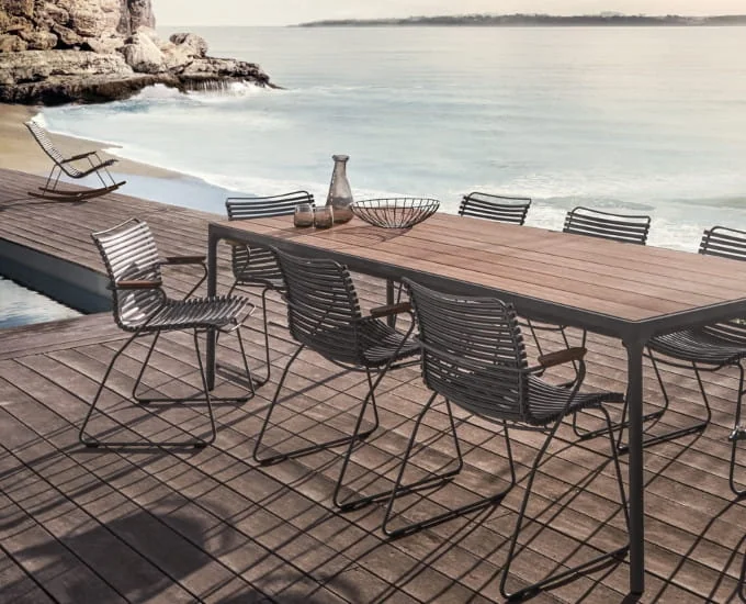 Prostokątny stół Four o wymiarach 210 x 90 cm z aluminiowym blatem z listew i metalowymi krzesłami na drewnianym tarasie nad oceanem.