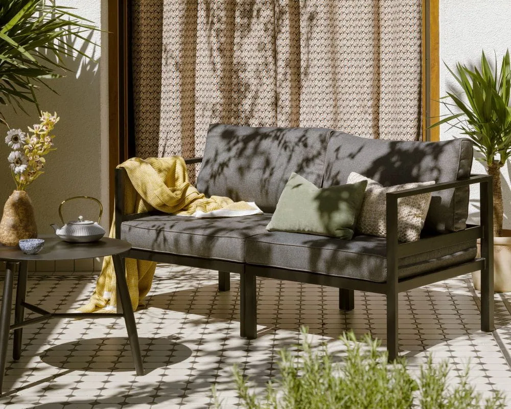 Modułowa sofa dwuosobowa Toulouse w kolorze ciemnoszarym z poduszkami, żółtym kocem i małym okrągłym stolikiem na tarasie.