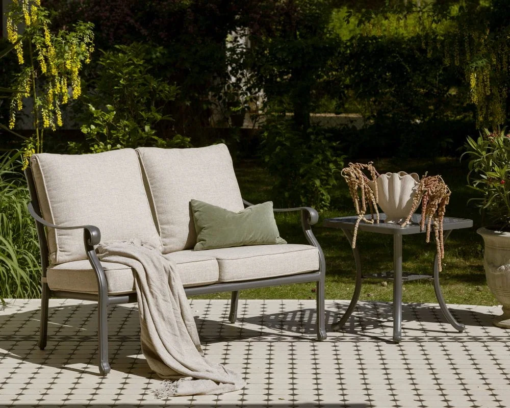 Dwuosobowa sofa w stylu prowansalskim „Vichy”, z beżowymi poduszkami, szarą metalową ramą, zielonym kocem i poduszką na tarasie.