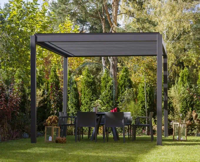 Aluminiowa pergola Saint Tropez z oświetleniem LED i dachem lamelowym nad zestawem jadalnianym na trawniku.
