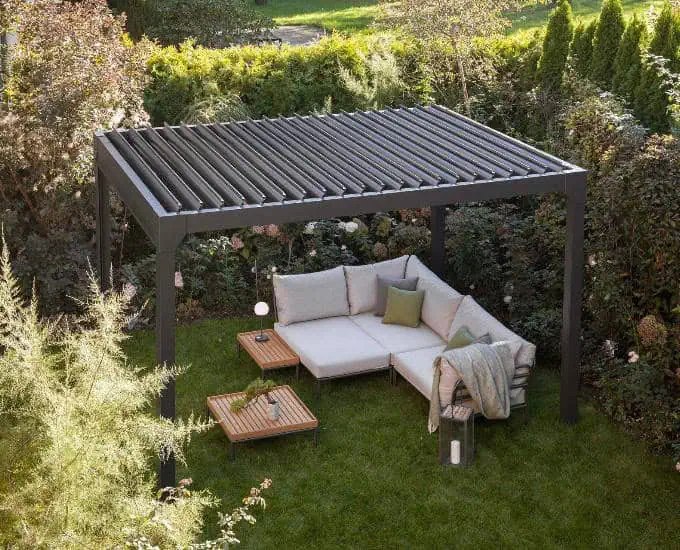 Pergola Monaco, aluminiowe zadaszenie sterowane elektrycznie z regulowanymi lamelami, narożnik i dwa drewniane stoliki na trawniku.